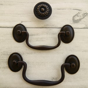 May include: Two antique bronze drawer pulls with a decorative oval shape and a curved handle. A matching antique bronze knob with a floral design is also shown.