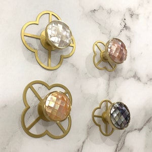 Può includere: Quattro ganci decorativi in metallo dorato con un design a quadrifoglio. Ogni gancio presenta un pomello rotondo, iridescente, in madreperla di un colore diverso: bianco, rosa, pesca e grigio.