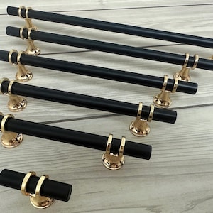 Può includere: Una collezione di maniglie per mobili nere con accenti color oro. Le maniglie sono cilindriche con anelli dorati e sono montate su basi color oro. L'hardware è disposto su una superficie a venature di legno chiaro.