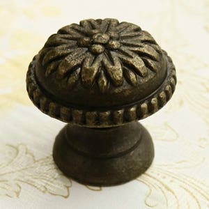 May include: A bronze-colored cabinet knob with a floral design. The knob has a round top and a cylindrical base.