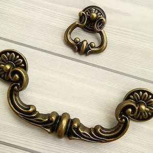May include: Two antique-style cabinet pulls with ornate floral designs. The larger pull is a D-shaped handle with a decorative knob on each end. The smaller pull is a ring-shaped handle with a decorative knob on top.