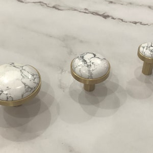 Op de afbeelding: Drie ronde kastknoppen van wit marmer en messing met een wit marmer bovenblad. De knoppen zijn in verschillende maten.