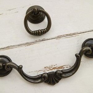 May include: Antique bronze cabinet hardware set. Includes a ring pull and a decorative handle with a curved design. The hardware features a rope-like border and ornate detailing, suitable for furniture restoration or new builds.
