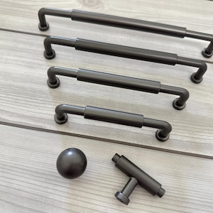 May include: A collection of matte black cabinet hardware including four varying lengths of cylindrical bar pulls, one spherical knob, and one T-shaped bar knob. Each piece features a smooth, uniform finish and simple, clean lines for attachment.