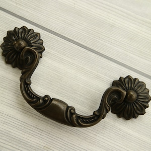 Könnte beinhalten: Antiker Bronze-Schubladengriff mit verzierten floralen Details. Der Griff hat eine geschwungene Form und zwei dekorative Rosetten.