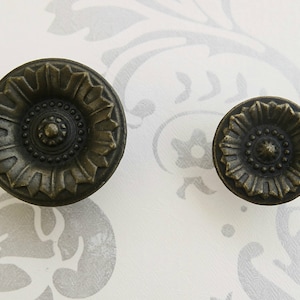Könnte beinhalten: Zwei antike Metall-Schrankknöpfe mit einem dekorativen Blumendesign. Die Knöpfe sind dunkelbronzefarben und haben eine erhabene Mitte mit einem kleineren Kreis in der Mitte.