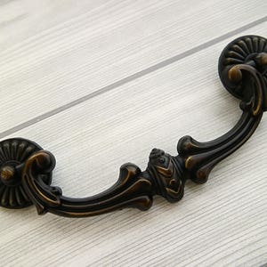May include: A black metal drawer pull with a decorative, ornate design. The pull has a curved shape and two round ends.