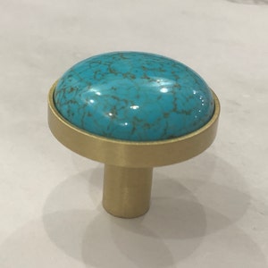 Może przedstawiać: Okrągły gałka do szafki w kolorze turkusowym i złotym. Gałka ma turkusowy kamień osadzony w złotej metalowej podstawie.