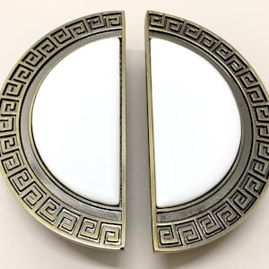 Könnte beinhalten: Zwei antike goldfarbene Schranktürgriffe mit einem weißen Keramikmittelteil. Die Griffe sind halbkreisförmig mit einem griechischen Schlüsselmuster am Rand.