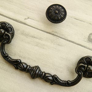 Può includere: Pomelli per cassetti e pomolo in bronzo scuro con un design ornato. Il pomello ha una maniglia curva con dettagli decorativi e accenti floreali. Il pomolo presenta un design floreale simile. Per mobili.