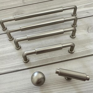 May include: A collection of brushed nickel cabinet hardware including four varying lengths of bar pulls, one round knob, and one T-bar knob. Each piece features a smooth, cylindrical design with rounded ends and a consistent metallic finish.
