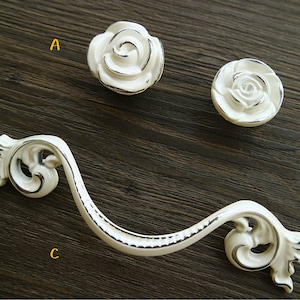 Puede incluir: Tres tiradores de cajón de cerámica blanca. Dos son redondos con un diseño de rosa en relieve. El tercero es un tirador curvo decorativo, adornado, con un acabado plateado.