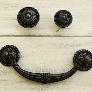 Puede incluir: Tirador de cajón de metal negro con detalles ornamentados. El tirador tiene una forma curva y dos pomos redondos en cada extremo.