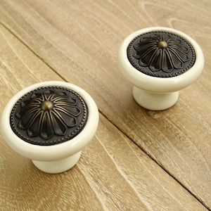 May include: Two white ceramic cabinet knobs with a bronze floral design. The knobs have a round base and a raised, circular top.