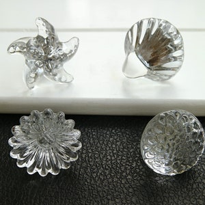 May include: Four clear glass cabinet knobs with different designs: a starfish, a seashell, a flower, and a textured circle.
