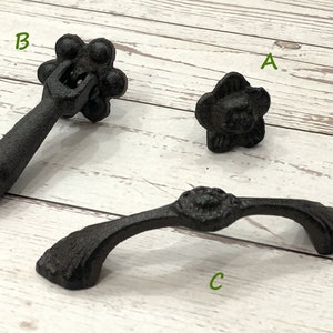 May include: Three black cast iron drawer pulls. The first pull is a small, round knob with a flower design. The second pull is a long, straight handle with a round knob at the end. The third pull is a curved handle with a round knob in the center.