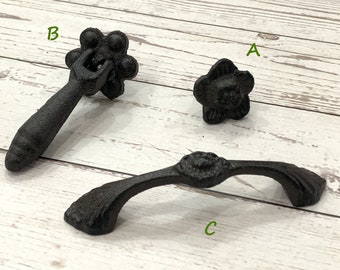 Könnte beinhalten: Drei schwarze Schubladengriffe aus Gusseisen. Der erste Griff ist ein kleiner, runder Knopf mit einem Blumendesign. Der zweite Griff ist ein langer, gerader Griff mit einem runden Knopf am Ende. Der dritte Griff ist ein gebogener Griff mit einem runden Knopf in der Mitte.