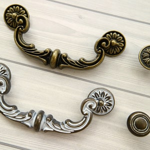 May include: Two antique-style drawer pulls and two matching knobs. The pulls have a curved handle design with floral accents and are finished in a bronze colour with white detailing. The knobs feature a floral design. The hardware is suitable for cabinets and furniture.