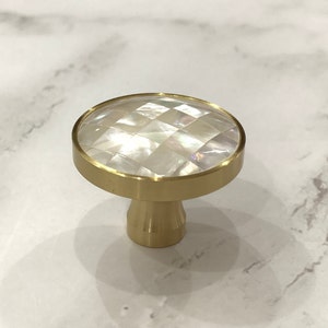 May include: A round, gold-toned cabinet knob with a white mother-of-pearl inlay. The inlay has a geometric pattern.
