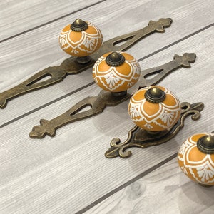 Könnte beinhalten: Dekorative Schrankbeschläge mit senfgelben Keramikknöpfen mit weißen Blumenmustern. Die Knöpfe sind auf antiken bronzefarbenen Griffen montiert, ideal für Möbel und Heimdekoration. Geeignet für Möbelprojekte.