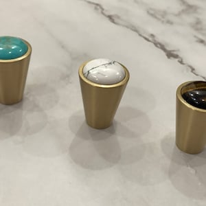 Może przedstawiać: Trzy złote gałki do szafek z różnokolorowymi kamiennymi wkładkami. Gałki są okrągłe i mają zwężającą się podstawę. Kamienie są turkusowe, białe i brązowe.