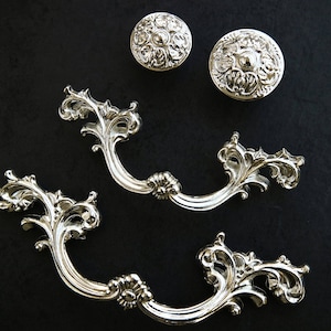 Könnte beinhalten: Drei silberfarbene Schrankknöpfe mit verzierten Blumenmustern. Die Knöpfe sind in verschiedenen Formen und Größen.
