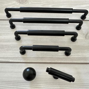 May include: A collection of matte black cabinet hardware including four varying lengths of bar pulls with cylindrical centres and rounded ends, one spherical knob, and one cylindrical T-bar knob, all featuring a smooth, uniform finish.
