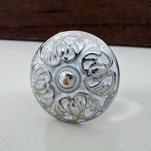Könnte beinhalten: Ein runder, weißer Schrankknopf mit silbernem Filigranmuster. Der Knopf hat einen zentralen silbernen Akzent und ein dekoratives, verziertes Muster. Die Hardware ist wahrscheinlich für Möbel oder Schränke.