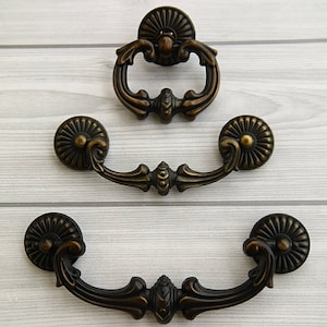 May include: Three antique brass cabinet pulls with ornate designs. The top pull is a ring pull with a decorative center. The two bottom pulls are D-shaped with decorative rosettes.