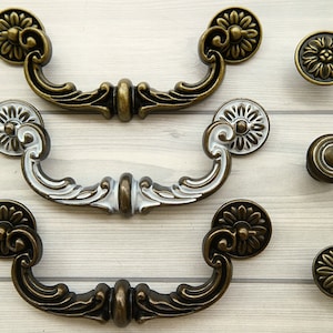 May include: Three ornate cabinet pulls with a floral design. Two are in a traditional pull style, one is a round knob. One pull is antique brass, one is antique silver, and the knob is antique brass.