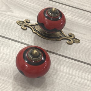 May include: Two red and black ceramic cabinet knobs with gold accents. The knobs are round and have a decorative design.