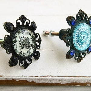 May include: Two ornate metal cabinet knobs with decorative glass insets. One knob has a black and white floral design, the other has a blue and white floral design. Both knobs have decorative metal accents.