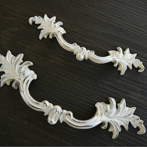 May include: Two ornate white metal cabinet pulls with a leaf design. The pulls have a silver finish.