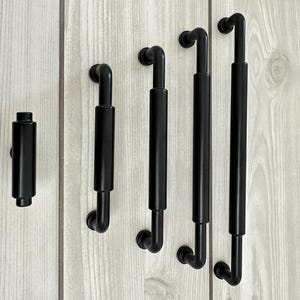 以下が含まれることがあります： マットブラック仕上げのキャビネット用ハードウェア5点セット。小さなノブ1個と、長さの異なる4本の円筒形バーハンドルで構成されており、それぞれ滑らかな質感と、家具やキャビネットに適した頑丈で直線的なデザインが特徴です。