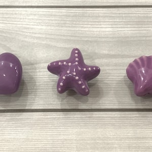 May include: Three purple ceramic drawer pulls in the shapes of a heart, a starfish, and a seashell.