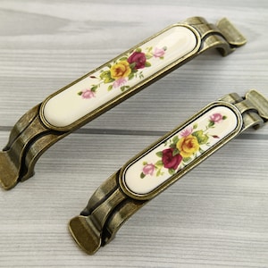 Może przedstawiać: Dwa antyczne złote gałki meblowe z białymi wkładkami ceramicznymi z kwiatowym wzorem z różowymi, czerwonymi i żółtymi różami.