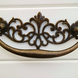 Op de afbeelding: Een decoratieve, sierlijke, messingkleurige ladegreep met een gebogen handvat en ingewikkelde krulwerk.
