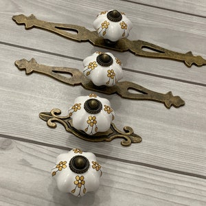 Może przedstawiać: Trzy białe ceramiczne gałki do szuflad z złotymi wzorami kwiatowymi i antycznymi mosiężnymi okuciami. Każda gałka ma ozdobną, zdobioną płytkę tylną.