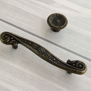 Może przedstawiać: Antyczny brązowy uchwyt do szafki z ozdobnym wzorem kwiatowym. Uchwyt ma zakrzywiony kształt i okrągły guzik na końcu.