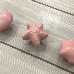 Könnte beinhalten: Drei rosafarbene Keramik-Schubladengriffe in Form eines Herzens, eines Seesterns und einer Muschel.