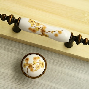 Może przedstawiać: Zestaw dwóch białych ceramicznych gałek do szafek z kwiatowym wzorem w odcieniach żółtego i brązowego. Gałki mają brązowe wykończenie i są przymocowane do drewnianej powierzchni.