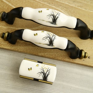 Puede incluir: Tres pomos de armario de cerámica blanca con bases de metal negro. Cada pomo presenta un delicado diseño floral con mariposas amarillas.