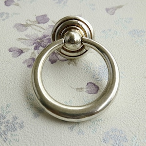 May include: A silver-toned cabinet pull with a ring handle. The pull has a circular base with concentric ridges and a small, rounded knob. The ring handle is attached to the base and is a simple, smooth circle. The hardware is likely for furniture.
