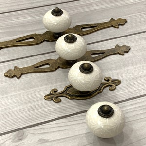 May include: Four white ceramic knobs with ornate designs and antique gold metal drawer pulls. The knobs are arranged in a row on a light wood surface.