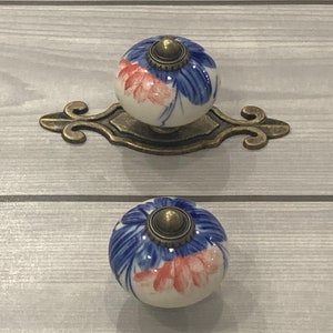 May include: Two white ceramic drawer knobs with blue and pink floral designs. The knobs have a brass finish and are attached to a decorative brass plate.
