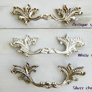 May include: Three ornate drawer pulls in different finishes: antique silver, white silver, and silver chrome. Each pull features a decorative, scroll-like design.
