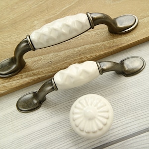 May include: Two white ceramic cabinet pulls with an antique silver finish. The pulls have a decorative, raised pattern. One pull is a knob and the other is a handle.