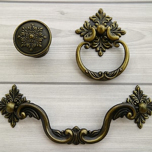 May include: Three antique-style cabinet knobs with ornate floral designs. The knobs are made of metal and have a brass finish. One knob is round, and the other two are shaped like a pull handle.