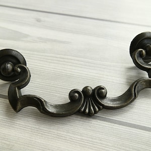 May include: Antique bronze-finished drawer pull with a decorative design. The pull features a curved handle with ornate detailing in the centre and circular end pieces. Suitable for cabinets and drawers.