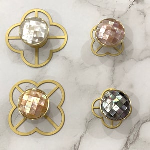 Pode incluir: Quatro botões de armário em tom dourado com um design decorativo de trevo. Cada botão apresenta uma incrustação redonda de madrepérola iridescente.